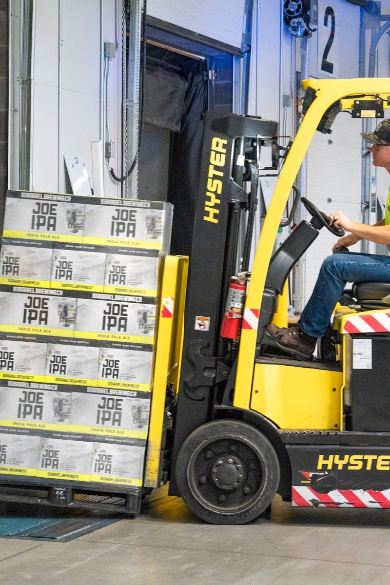 A worker drives a Hyster forklift moving Joe IPA boxes in a warehouse.